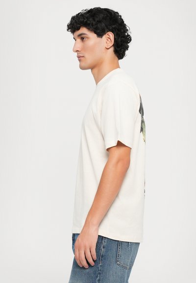 T-shirt color crema con maniche corte e colletto tondo, con un design grafico colorato sul retro, indossata con jeans in denim blu.