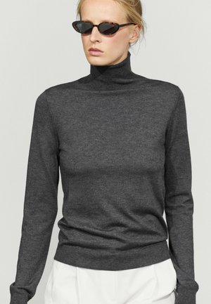 Vrouw met zwarte cat-eye zonnebril, donkergrijze coltrui en witte broek tegen een lichtgrijze achtergrond.