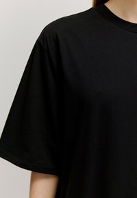 Schwarzes Baumwoll-T-Shirt mit rundem Ausschnitt und kurzen, weiten Ärmeln. Der Stoff wirkt weich und glatt, ohne sichtbare Muster oder Akzente.