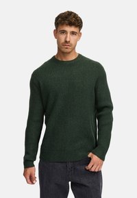 Jersey de punto de canalé verde con cuello redondo, mangas largas y un corte relajado, combinado con jeans oscuros.