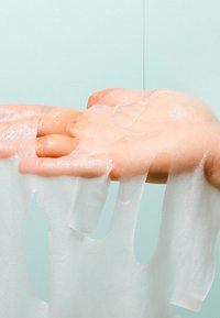Hand with gel-like substance dripping from fingers and a clear liquid pouring onto the back of the hand against a light background.