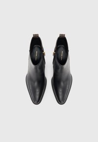 Stivali neri in pelle fino alla caviglia con punta affusolata, chiusura con zip e dettagli in tessuto marrone sul retro. Texture liscia, design minimalista.