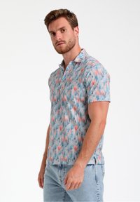 Camisa polo de manga corta en azul claro con un patrón abstracto colorido de formas rosas y verdes, con un cuello clásico y tapeta de botones.