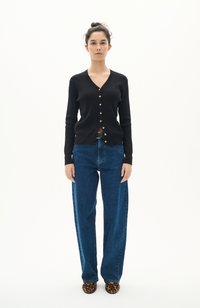 Svart ribbad cardigan med guldfärgad knappar, kombinerad med högmidjade blå denimjeans. Modellen bär klackar i leopardmönster. Minimalistisk bakgrund.