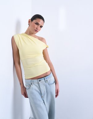 Γυναίκα ακουμπισμένη σε λευκό τοίχο, φορώντας κίτρινο τοπ με έναν ώμο και ανοιχτόχρωμο τζιν ψηλόμεσο, κοιτώντας την κάμερα.