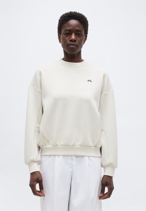 Άτομο που φοράει απλό off-white φούτερ με μικρό μαύρο λογότυπο στο στήθος και άσπρο παντελόνι, στέκεται μπροστά σε απλό φωτεινό φόντο.