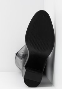 Bottine en cuir noir avec fermeture éclair latérale, montrant la semelle en taille 37, positionnée à la verticale contre un fond d'angle blanc.