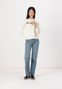Witte lange mouwen trui met een luipaardprint ontwerp en groene accenten, gecombineerd met lichtblauwe jeans en zwarte sneakers.