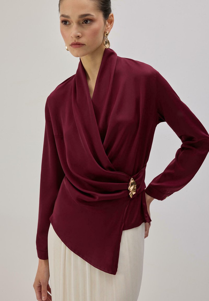 Femme portant un chemisier en satin bordeaux avec un devant drapé et une boucle dorée, associé à une jupe plissée blanche, debout devant un fond uni.