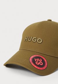 Olivovo zelená čiapka s vyšitým logom "HUGO". Pevná tkanina, zakrivený šilt a červený kruhový označník s detailmi značky na boku.