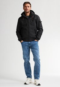 Chaqueta negra con capucha, cierre de cremallera, cordones ajustables, bolsillos laterales y puños acanalados, combinada con jeans de mezclilla azul claro y zapatillas blancas.