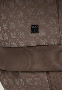 Braun gemusteter Stoff mit floralem Design und glatter Textur. Ein kleines schwarzes metallisches Logosquare ist am Kleidungsstück befestigt.