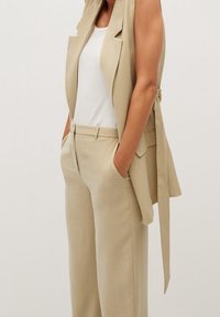 Beżowa kamizelka o dopasowanym kroju, z długim fasonem, szerokimi klapami i wiązanym akcentem w talii, zestawiona z pasującymi spodniami o wysokim stanie. Gładka tekstura materiału.