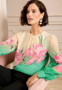Blusa floreale con un design trasparente crema e verde, decorata con motivi di fiori rosa, con maniche lunghe e scollatura arricciata.