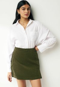 Cămașă albă cu mâneci lungi și un buzunar, asociată cu o fustă mini din corduroy verde închis, cu dungi verticale.