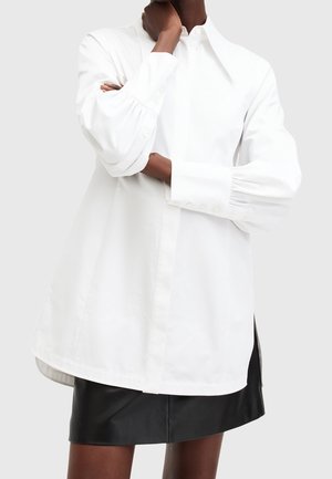 Woman wearing a white long-sleeve oversized shirt with puffed cuffs and a black leather mini skirt against a plain background.