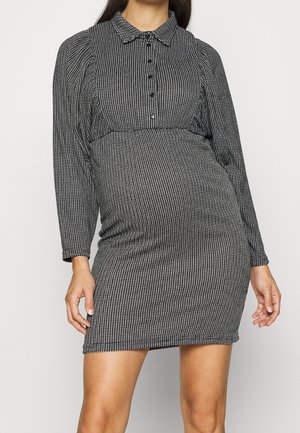 Femme portant une robe à manches longues à motif noir et blanc avec des boutons sur la poitrine et un col, debout devant un fond uni.