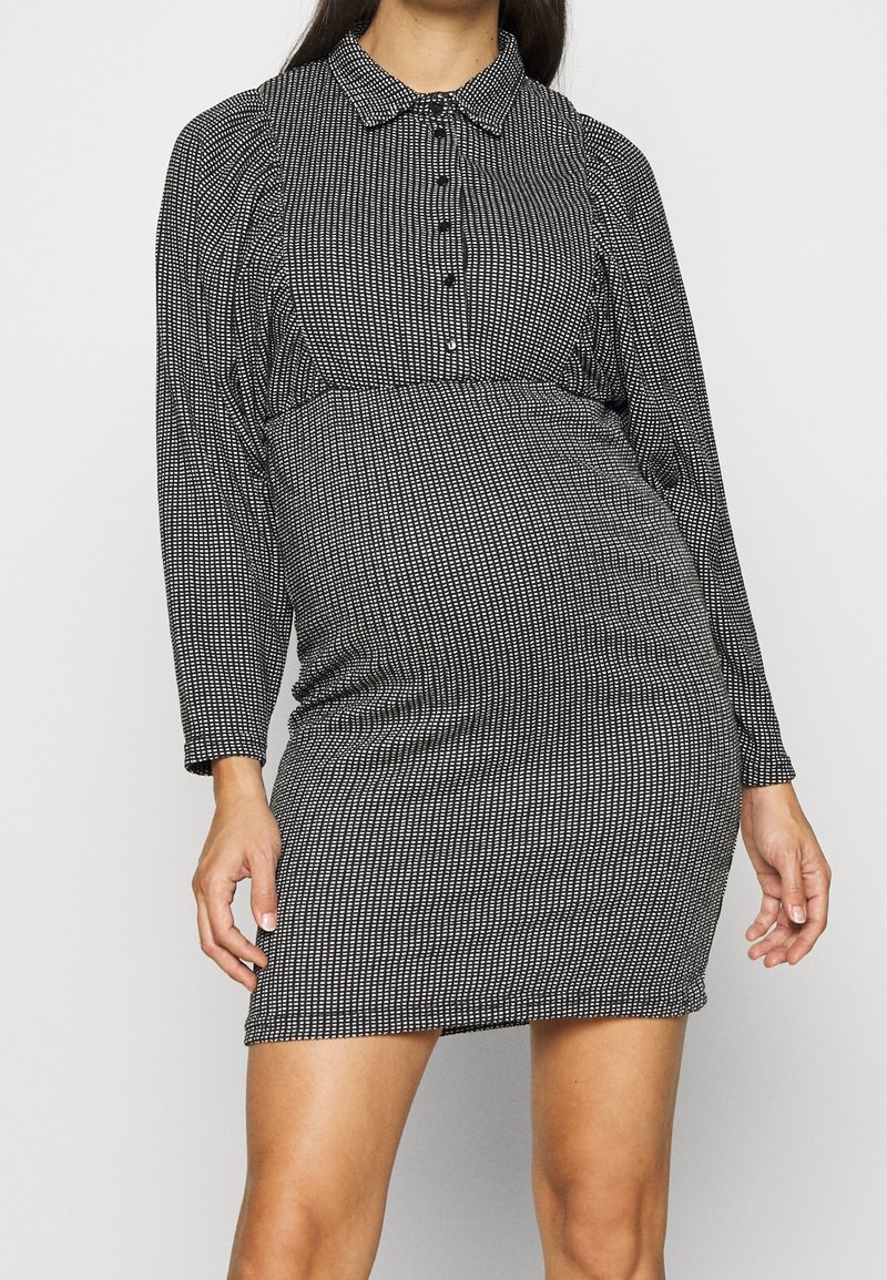 Femme portant une robe à manches longues à motif noir et blanc avec des boutons sur la poitrine et un col, debout devant un fond uni.