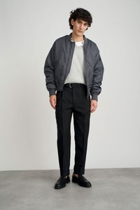 Grijze bomberjack met een gequilt textuur over een lichtgrijze trui, gecombineerd met zwarte op maat gemaakte broek en zwarte instapschoenen.
