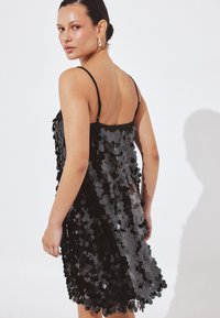 Nainen, jolla on tumma tukka matalalla nutturalla, pukeutunut hihattomaan mustaan mekkoon, joka on koristeltu suurilla kiiltävillä paljeteilla, katsoo yli olkapäänsä valkoista taustaa vasten.