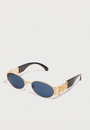 Lunettes de soleil ovales à verres bleu foncé avec montures en métal doré et détails décoratifs dorés sur des branches en écailles de tortue foncées.