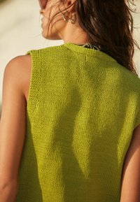 Kobieta z długimi ciemnymi włosami, ubrana w zielony bezrękawnik z dzianiny i koralikowe kolczyki w kształcie kół, pokazana z tyłu i w profilu.