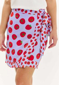 Zavinovací sukňa z svetlo levanduľovej textílie, ktorá má živý potlač červených paradajok s zelenými akcentmi. Uviazaná v pásu s detailom mašle.