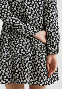 Schwarzes Kleid aus Stoff mit einem weißen Gänseblümchenmuster, langen Ärmeln, gerafftem Rock und lässiger Passform. Die Textur wirkt leicht und fließend.