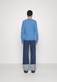 Kvinna med hår i en knut, klädd i en blå långärmad topp och mörka vida jeans med breda vikta fållar, stående mot en slät vit bakgrund.