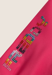 Fuksia kangas, jossa on sileä pinta ja monivärisiä strassikoristeita, jotka muodostavat sanan "VIBRANT" eri kokoisina ja muotoisina.