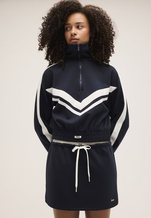 Mujer con cabello rizado que lleva una sudadera azul marino y blanca con cremallera y una falda a juego con cintura ajustable con cordón, de pie contra un fondo liso.