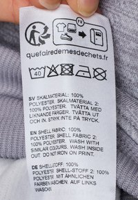 Etichetta con istruzioni per la cura in più lingue. Tessuto bianco con stampa nera, inclusi icone di lavanderia e testi sui materiali e il lavaggio.