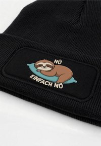 Black ribbed beanie hat featuring a fabric patch with a cartoon sloth resting on a teal pillow and the text “NÖ EINFACH NÖ” beneath.
