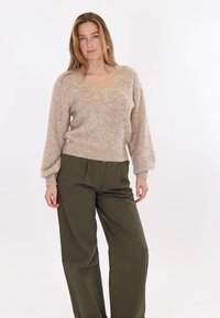 Donna in piedi con postura rilassata, indossa un maglione peloso beige e pantaloni ampi verde oliva, su sfondo bianco semplice.