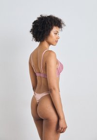 Rózsaszín, textúrált fehérnemű szett, amely egy vékony pántos bralette-ből és egy hozzá illő tanga alsóból áll, sima selyem és csipke részletekkel.
