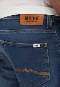Džinsai su rudu odiniu žymeniu "MUSTANG True Denim", mėlyna medžiaga, geltonos siūlės ir logotipas galiniame kišenėje.