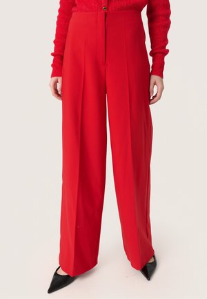 Femme portant un pantalon rouge taille haute à jambes larges, un haut rouge à manches longues et des escarpins noirs à bout pointu, debout devant un fond uni.