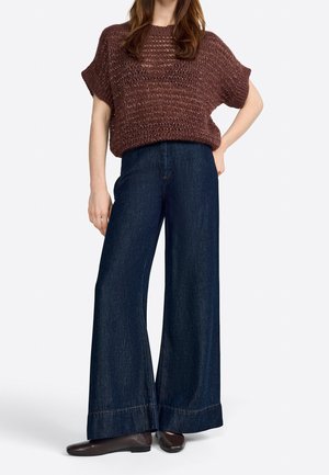 Woman wearing a loose brown knit short-sleeve sweater, wide-leg dark blue jeans, and dark brown flat shoes standing with one hand on hip.