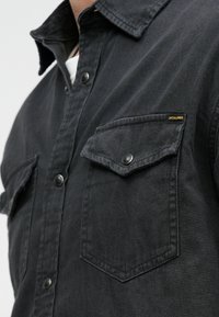 Primo piano di una camicia button-up grigia scura con tasche a soffietto sul petto e l'etichetta "JACK & JONES" ben visibile sulla tasca destra.