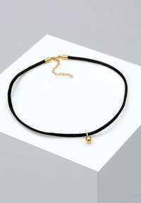 Collier ras du cou en corde noire avec fermoir doré, chaîne réglable, et petit pendentif sphérique doré reposant sur une surface blanche.