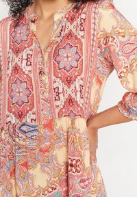 En flytande klänning med en blandning av intrikata mönster i röd, rosa och krämfärgad paisley, med knappslå framtill och ärmar som slutar vid tre fjärdedelar.