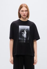 Μαύρο βαμβακερό μπλουζάκι με λευκό γραφικό τύπωμα χορευτή στο κέντρο. Κοντά μανίκια και άνετη εφαρμογή, με στρογγυλή λαιμόκοψη.