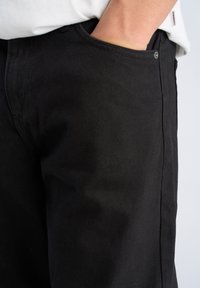 Pantaloni in denim nero con una texture liscia, dotati di tasca frontale e chiusura con bottone. Il tessuto presenta un motivo diagonale sottile.