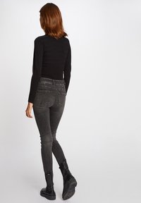 Mujer con cabello castaño rojizo hasta los hombros, vestida con una camiseta negra de manga larga, jeans oscuros ajustados y botas negras de tobillo, alejándose.