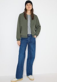 Chaqueta bomber verde oliva, camiseta gris, vaqueros acampanados azules y zapatillas blancas. Modelo con un gorro de punto negro, de pie contra un fondo blanco.