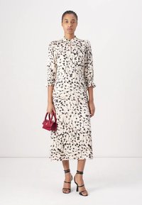 Cremefarbenes Langarmkleid mit schwarzem Blattmuster, gerafften Manschetten und Knopfleiste. Das Model hält eine kleine bordeauxfarbene Handtasche und trägt schwarze Riemchenheels.