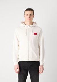 Felpa bianca con zip realizzata in tessuto morbido, con una patch logo rossa sul petto, tasche frontali e cappuccio con cordoncino. Indossata con jeans scuri.