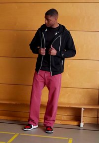 Sudadera con cremallera negra, camiseta negra, pantalones rojos y zapatillas negras/rojas. Tejido suave, corte relajado y acentos de cremallera blanca en contraste.