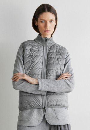 Kobieta o krótkich brązowych włosach, ubrana w szarą pikowaną kamizelkę bez rękawów na szarym długim rękawie, ze skrzyżowanymi ramionami, na białym tle.