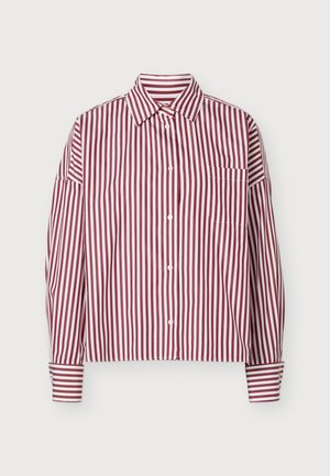 Femme portant une chemise rayée verticale rouge et blanche rentrée dans une longue jupe plissée bordeaux avec des bottes noires montantes jusqu'aux genoux.
